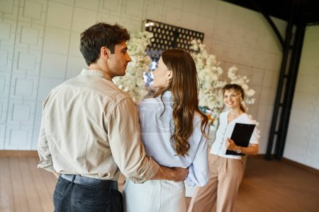 couple in love looking at each other near event coordinator in banquet hall with floral decoration