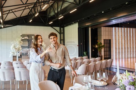 amazed woman pointing with hand near festive table and boyfriend in event hall with wedding decor