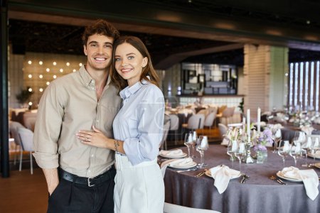 Foto de Feliz pareja enamorada mirando a la cámara en salón de banquetes con mesas de fiesta decoradas, día especial - Imagen libre de derechos