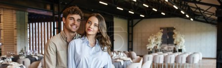 Photo for Cheerful couple looking at camera in modern and spacious banquet hall with wedding decor, banner - Royalty Free Image