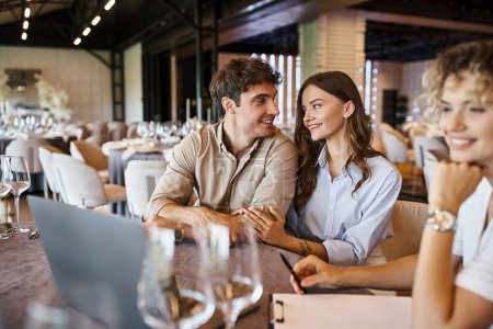 happy couple talking near blurred event manager near laptop on festive table in banquet hall