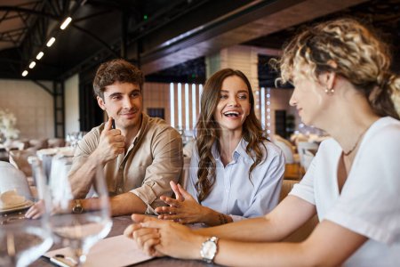 cheerful couple discussing wedding preparation with event manager at festive table in banquet hall