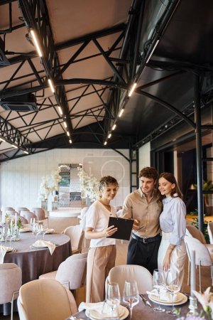 Photo for Joyful couple looking at contract on clipboard near banquet manager in decorated event hall - Royalty Free Image