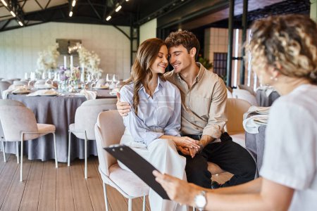 happy enamored couple sitting with closed eyes near event manager with clipboard in wedding venue