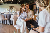 happy couple looking at blurred event manager while sitting at festive table in wedding hall t-shirt #675850590