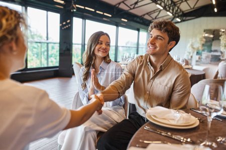 Lächelnder Mann beim Händeschütteln mit Eventmanager nahe überglücklicher Freundin in moderner Hochzeitslocation