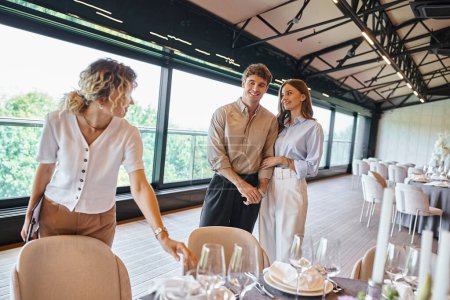 fröhliches verliebtes Paar neben Eventmanager und Tischen mit festlichem Rahmen in modernem Bankettsaal