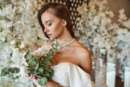 elegant woman in white wedding dress posing with bridal bouquet near decoration with white flowers