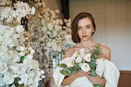 Photo for Elegant woman in white wedding dress with bridal bouquet looking at camera near festive floral decor - Royalty Free Image