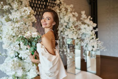 mujer joven feliz en vestido de novia posando con ramo de novia cerca de la decoración floral en la sala de eventos