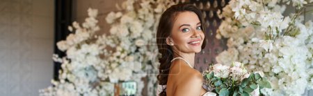 smiling bride in white wedding dress looking at camera in event hall with floral decor, banner