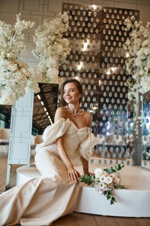 fröhliche junge Braut im weißen Hochzeitskleid sitzt neben weißem Blumendekor in der Festhalle