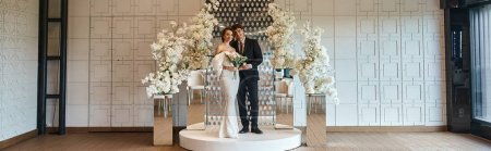 full length of elegant newlyweds posing in event hall decorated with white blooming flowers, banner