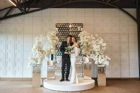 full length of romantic newlywed couple posing in event hall decorated with white blooming flowers