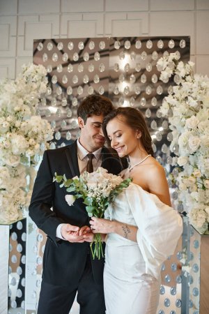 couple joyeux en tenue de mariage avec bouquet nuptial dans la salle des événements décorée de fleurs blanches