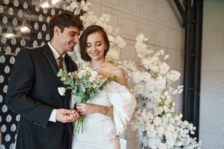 happy groom holding hand of charming bride with bridal bouquet near floral decor in event hall