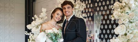 happy and elegant newlyweds smiling at camera in wedding hall decorated with white flowers, banner