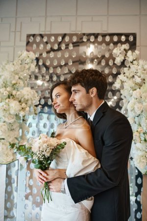 elegant groom embracing charming woman with bridal bouquet near white floral decor in banquet hall