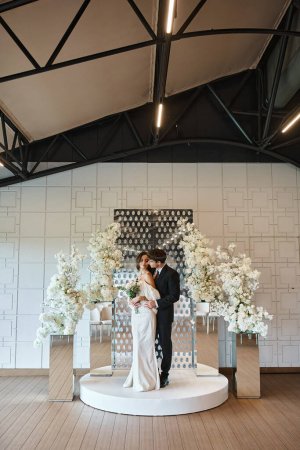 full length of groom embracing charming woman with bridal bouquet near floral decor in banquet hall