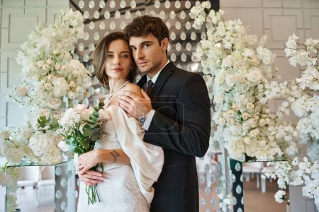young and elegant newlyweds looking at camera near white floral composition in wedding venue