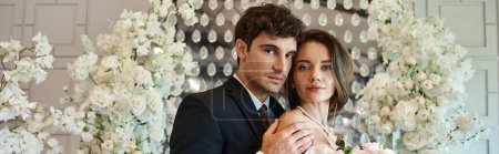 élégants jeunes mariés regardant la caméra près de composition florale blanche dans le lieu de mariage, bannière