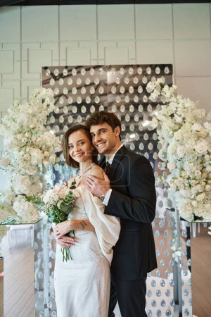 cheerful and elegant newlyweds smiling at camera near white floral composition in modern event hall