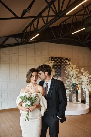 charming bride in white wedding dress near groom in black suit in event hall with floral decor