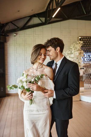 glückliche und charmante Braut mit Brautstrauß lächelnd neben elegantem Bräutigam in dekorierter Veranstaltungshalle