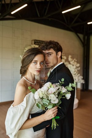 elegant and romantic newlyweds looking at camera near wedding bouquet in modern banquet hall