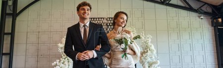 happy newlywed couple in wedding attire walking in modern event hall with white floral decor, banner