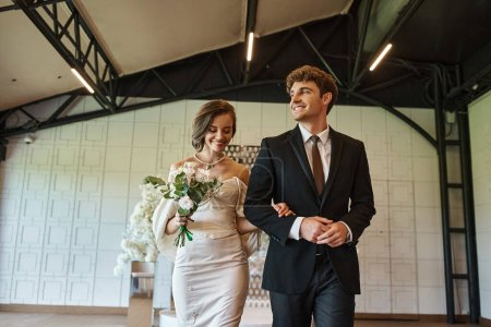 happy bride in white dress and groom in black suit smiling in modern decorated wedding venue