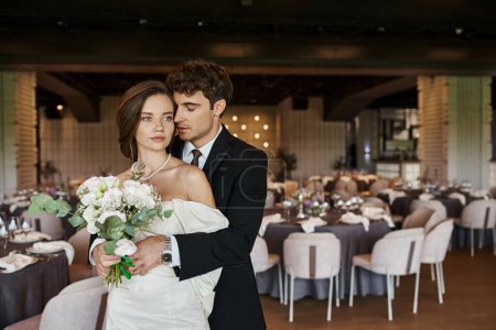 elegant man with closed eyes embracing young bride with wedding bouquet in modern event hall