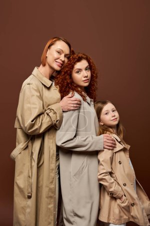 three generations, happy redhead women and cute girl in trench coats posing on brown background