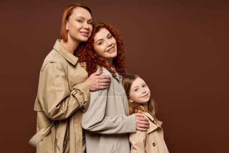 drei Generationen, fröhliche rothaarige Frauen und süße Mädchen in Trenchcoats posieren auf braunem Hintergrund