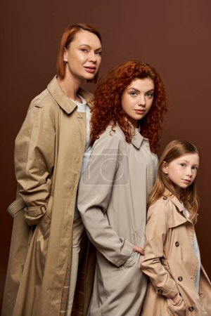 three generations, beautiful redhead women and girl in autumnal coats posing on brown background