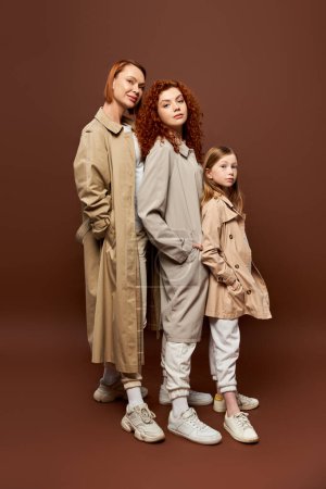 drei Generationen, rothaarige Frauen und Mädchen posieren in Mänteln vor braunem Hintergrund, herbstliche Kleidung