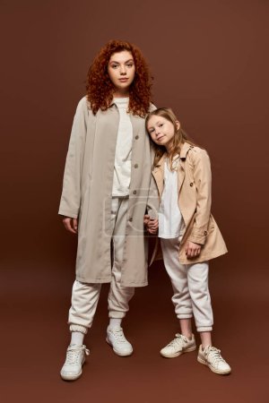 two generations, curly mother holding hands with daughter and standing in coats on brown backdrop