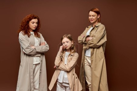 drei Generationen, Frauen in Mänteln, die vor braunem Hintergrund posieren und das Kind betrachten, rothaarige Familie