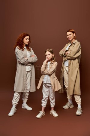 Photo for Three generations, women in coats posing and looking at girl on brown backdrop, redhead family - Royalty Free Image