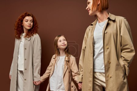 three generations, women in coats holding hands with happy girl on brown backdrop, redhead family