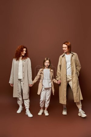Foto de Generación femenina, alegre familia pelirroja en abrigos tomados de la mano sobre fondo marrón, mujeres y niñas - Imagen libre de derechos