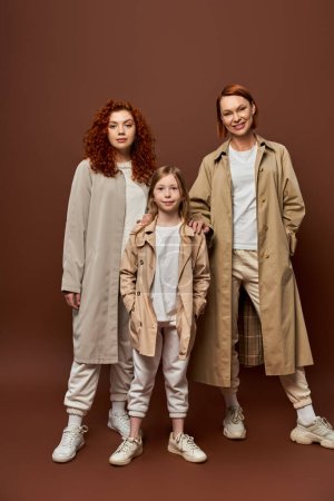 Téléchargez les photos : Génération féminine, rousse famille debout ensemble dans des vêtements de dessus sur fond brun, les femmes et les filles - en image libre de droit