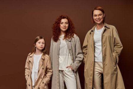 female generation, family with red hair posing with hands in pockets in outerwear on brown backdrop