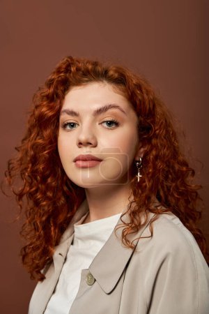 portrait of curly redhead woman with natural makeup looking at camera on brown background, autumn