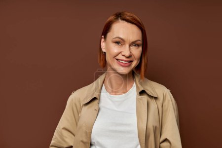 happy redhead woman in beige trench coat looking at camera on brown backdrop, autumn fashion