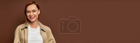 heureuse rousse femme en trench coat beige regardant la caméra sur fond brun, mode automne, bannière