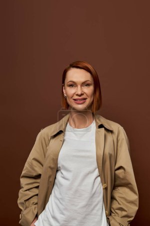 joyful redhead woman in beige trench coat looking at camera on brown backdrop, autumn fashion