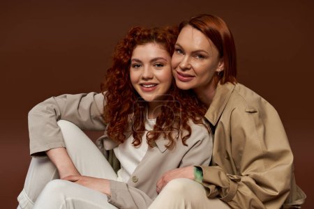 two generations, cheerful women with red hair posing in trendy autumn attire on brown background