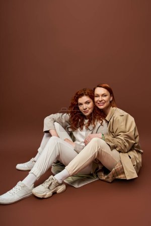 two generations, positive women with red hair posing in autumn trench coats on brown background