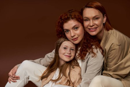 drei Generationen von Frauen, glückliche rothaarige Familie, die in die Kamera schaut und auf braunem Hintergrund lächelt